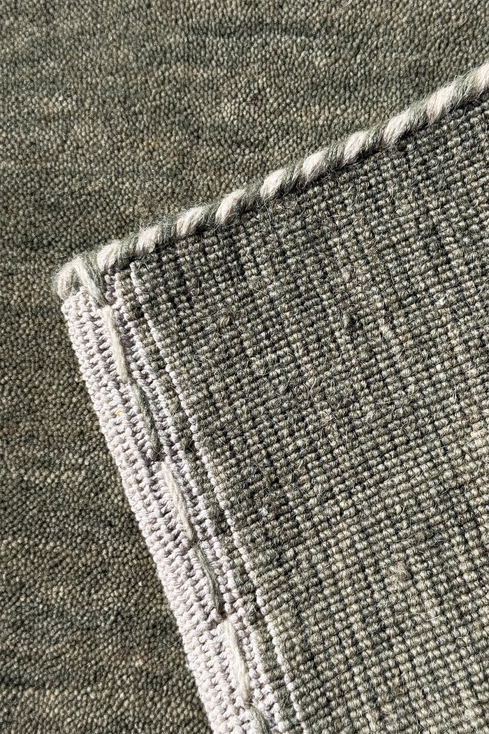 Close-up of a textured sage woven rug corner with a twisted light gray rope border.