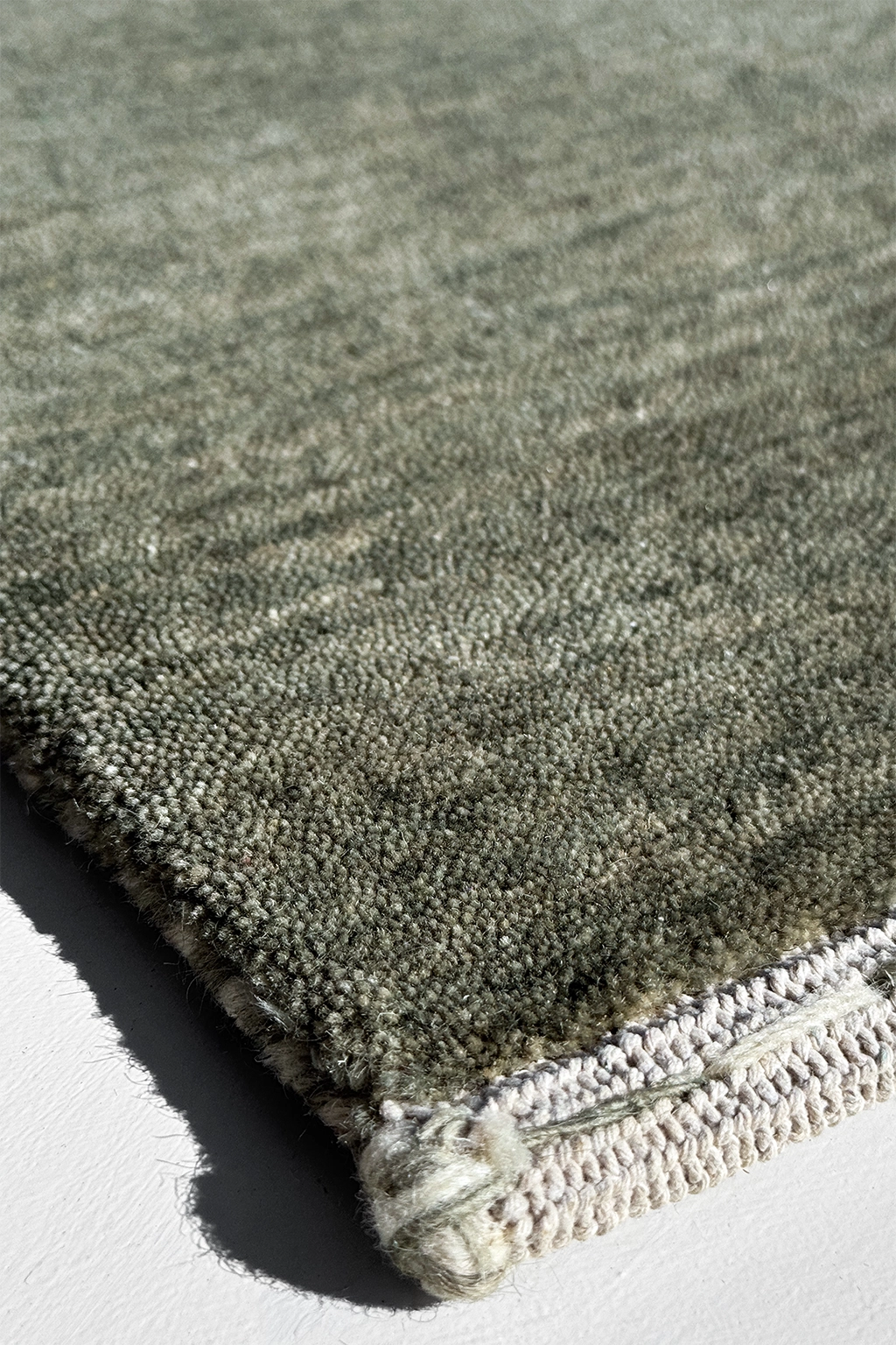 Close-up of a textured area rug in a muted sage green-gray color, showing the pile texture and a cream-colored fringed edge on a white surface.