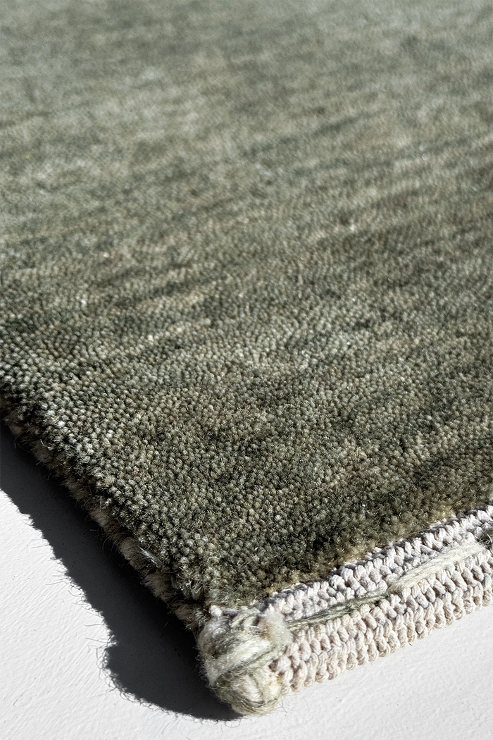 Close-up of a textured area rug in a muted sage green-gray color, showing the pile texture and a cream-colored fringed edge on a white surface.
