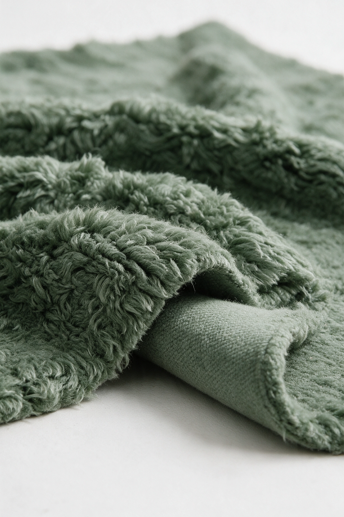 A close-up of a sage green plush shag rug with a textured, looped surface and a folded over edge showing the plush pile, set against a clean white background.