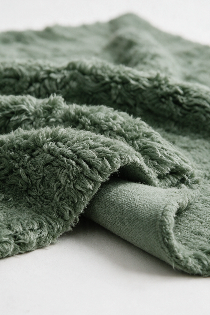 A close-up of a sage green plush shag rug with a textured, looped surface and a folded over edge showing the plush pile, set against a clean white background.