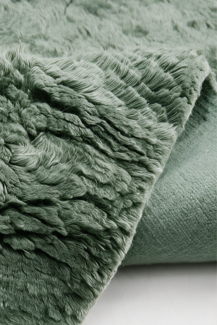 A close-up of a soft, plush wool rug in a muted sage green color, folded over to show both the shaggy texture and a smoother backing.