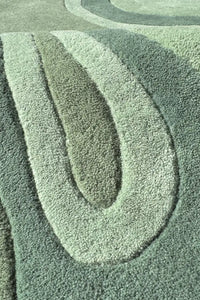 A close-up of a textured area rug with a modern wavy, concentric pattern in muted sage green, gray, and light gray tones.