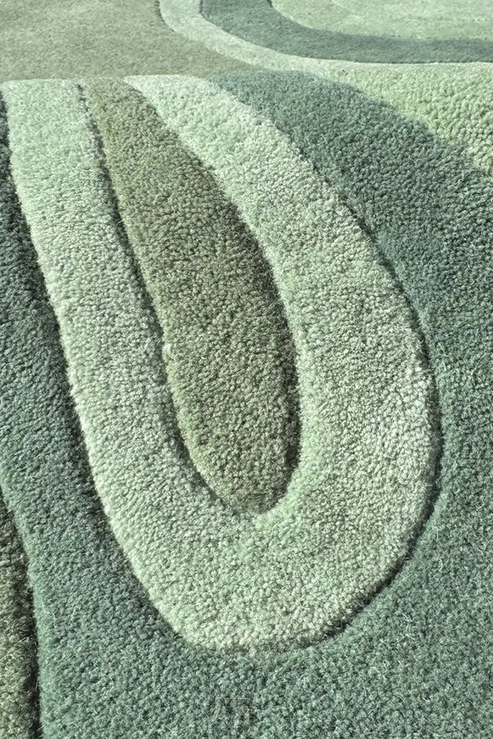 A close-up of a textured area rug with a modern wavy, concentric pattern in muted sage green, gray, and light gray tones.