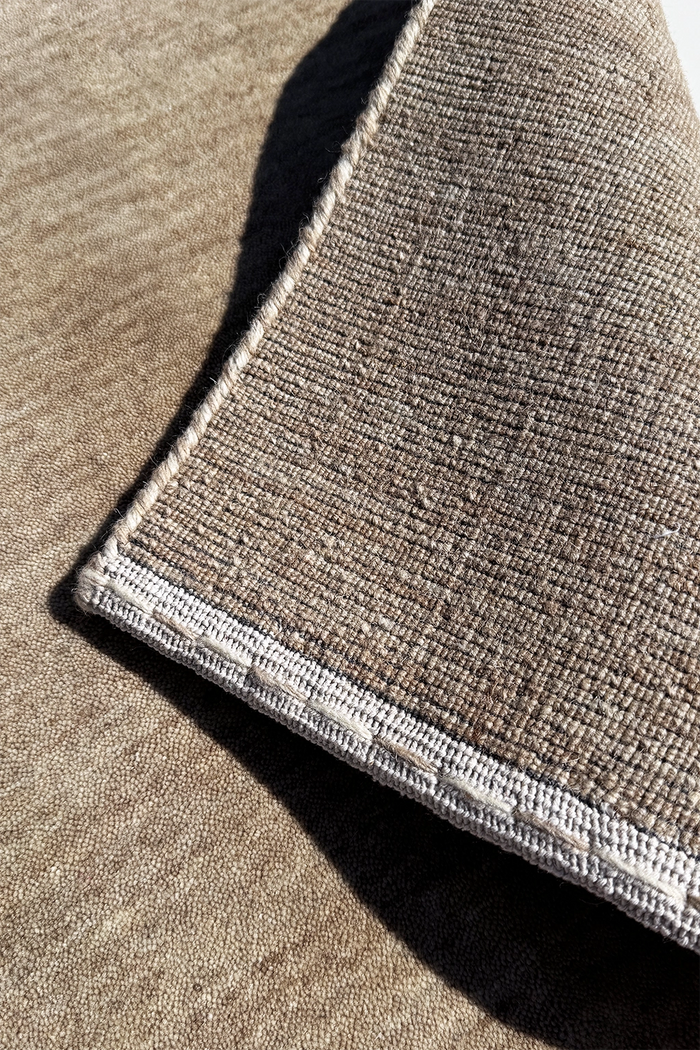 Close-up of a neutral runner rug showing a taupe-brown pile with a contrasting light beige stitched edge stone texture 