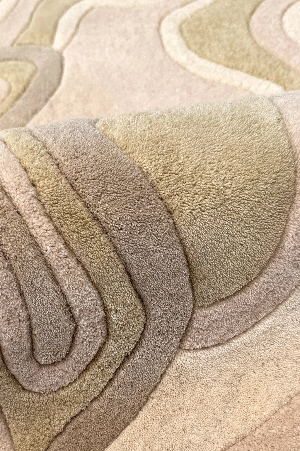A close-up of a plush, layered area rug with a wavy, topographic pattern in neutral tones of tan, beige, taupe, and light brown.