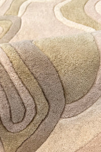 A close-up of a plush, layered area rug with a wavy, topographic pattern in neutral tones of tan, beige, taupe, and light brown.