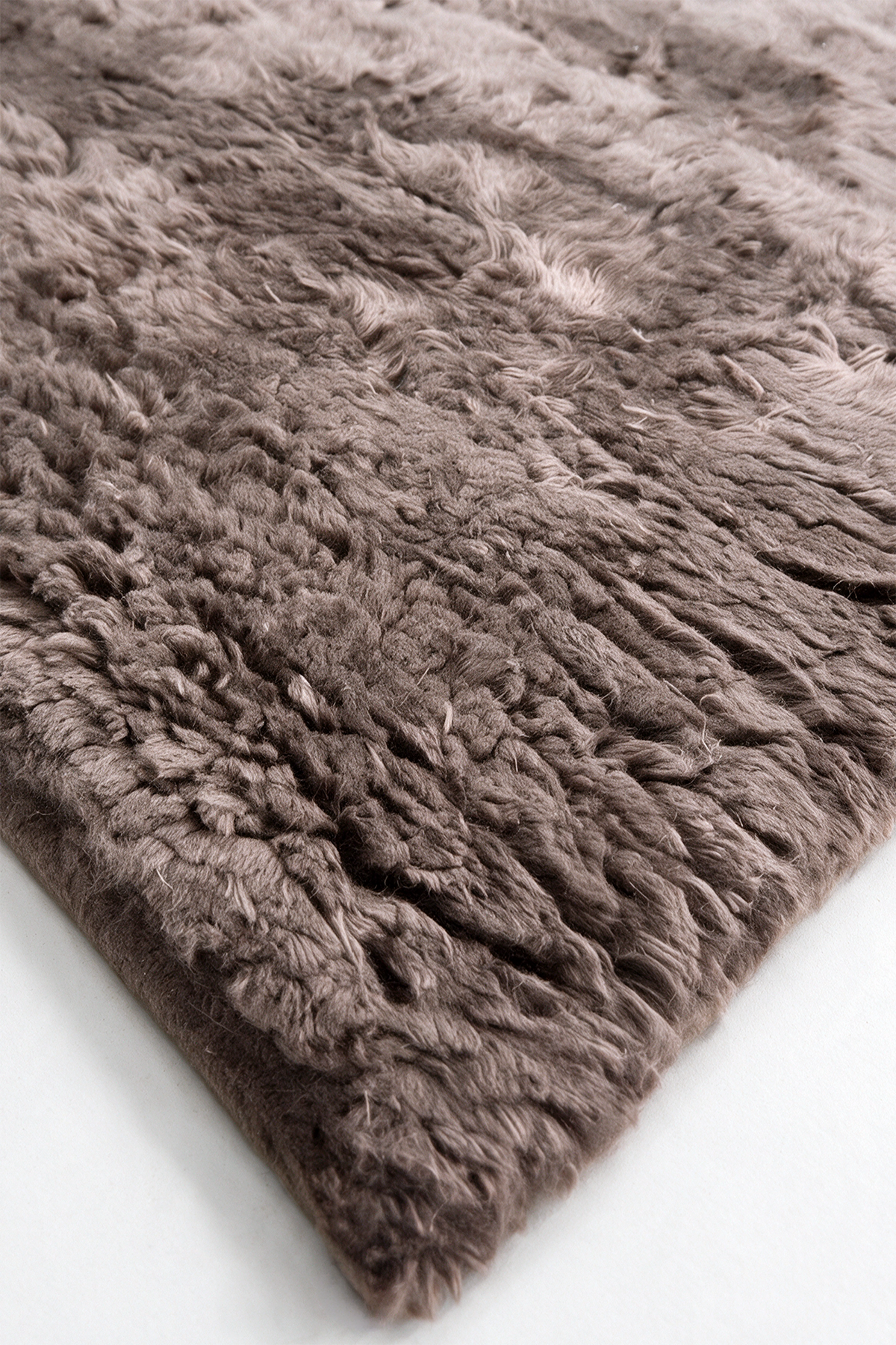 A close-up of a shaggy, long-pile rug in a taupe-brown color, showing its thick texture and looped fibers, with one corner folded over on a white surface.