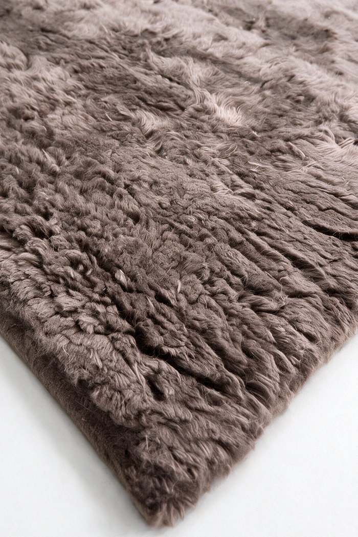 A close-up of a shaggy, long-pile rug in a taupe-brown color, showing its thick texture and looped fibers, with one corner folded over on a white surface.