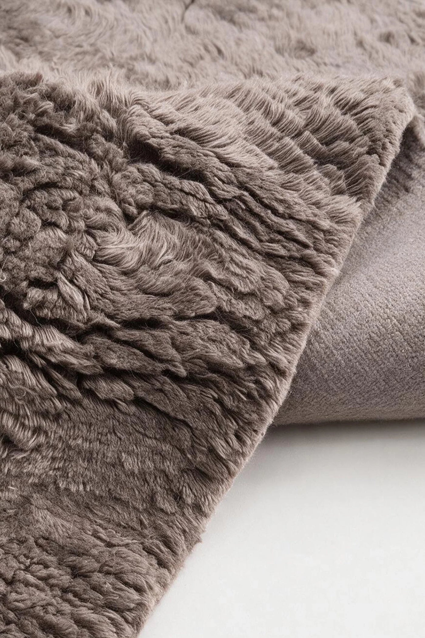 A close-up of a plush, textured wool area rug in a warm taupe-brown color, showing its shaggy pile and folded corner on a light neutral surface.