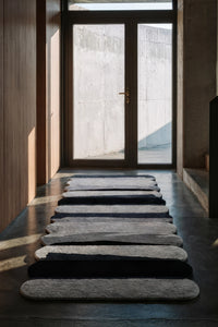 A modern, irregularly shaped runner rug in shades of gray, black, silver and white, arranged in a row on a dark concrete floor leading towards a glass door in a minimalist interior lifestyle