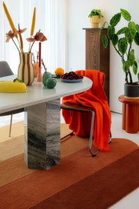 A modern dining room scene featuring a round white table with a gray marble pedestal base, styled with a bright orange throw blanket draped over a chair, a striped vase with dried flowers, and a striped terracotta rug.