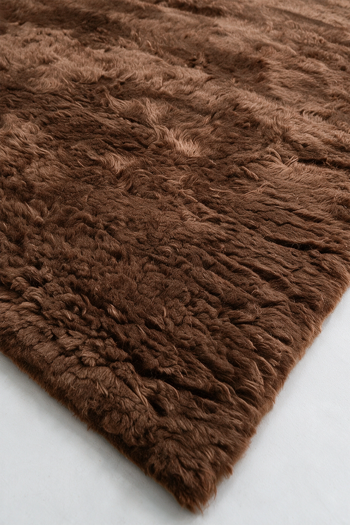 A close-up of a brown shaggy wool rug showing its thick, textured pile and corner detail against a light gray background umber