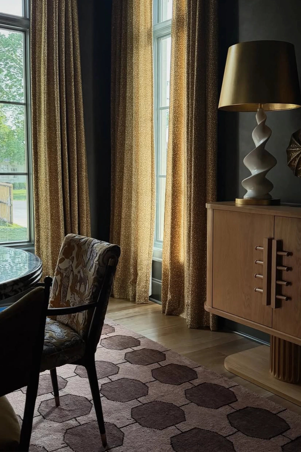 A styled interior corner with floor-length textured curtains in warm beige and gold tones, a wooden cabinet with a sculptural white lamp, a patterned dining chair, and a geometric rug on a light wood floor.