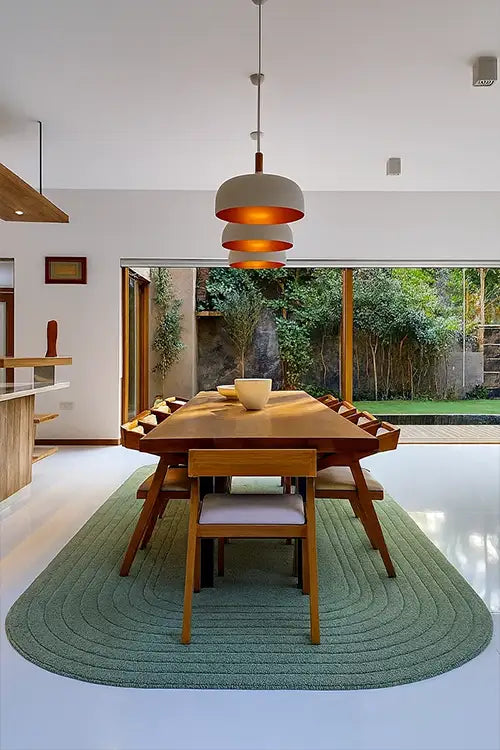 Modern dining area with wooden table and chairs on a green oval rug, pendant light, and large windows.