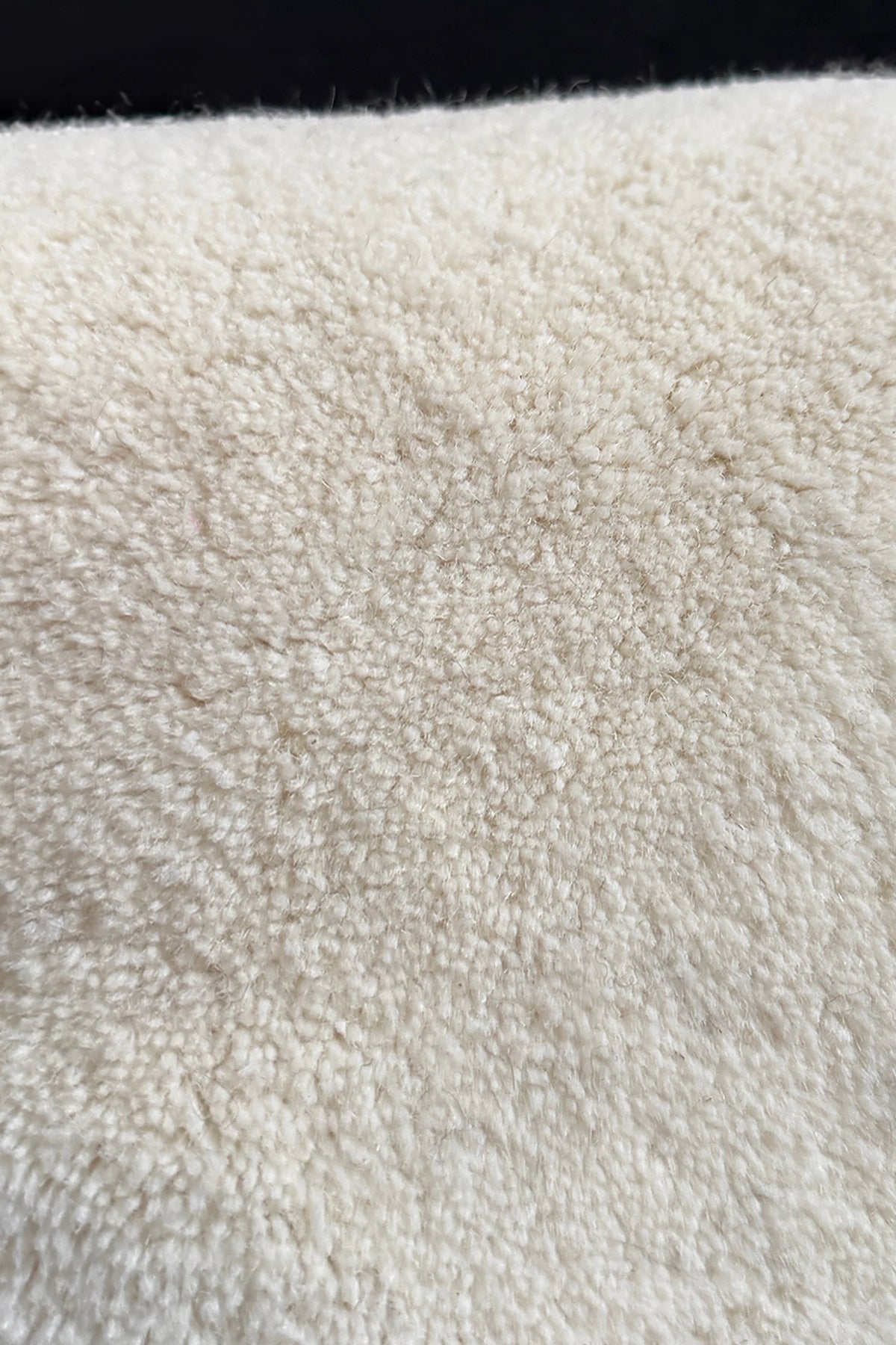 A close-up texture shot of a soft, plush wool rug in a warm off-white or cream color, with a dark black background visible at the top edge.