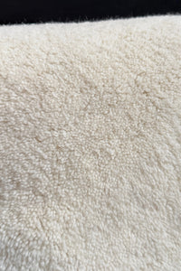 A close-up texture shot of a soft, plush wool rug in a warm off-white or cream color, with a dark black background visible at the top edge.