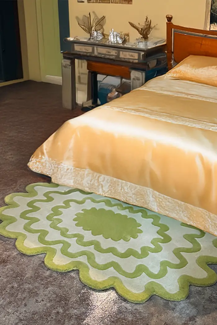 Burst Oval Rug in green with scalloped edges beside a gold satin bedspread in a vintage-inspired bedroom.
