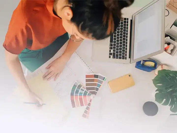 An interior designer working on a floor plan sketch with a set of color swatches, a laptop, and a phone on a bright workspace.