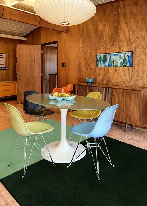 A stylish mid-century modern dining space with a two-tone green area rug, a tulip dining table, and colorful molded Eames-style chairs against wood-paneled walls.