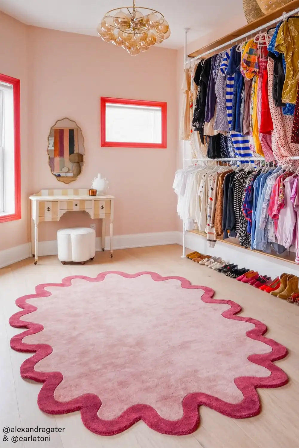 A stylish dressing room featuring a pink and red scalloped-edge rug as the centerpiece. The space has soft pink walls with bold coral window frames, an elegant vintage-style vanity, and an open closet showcasing a curated collection of colorful clothing and shoes. 