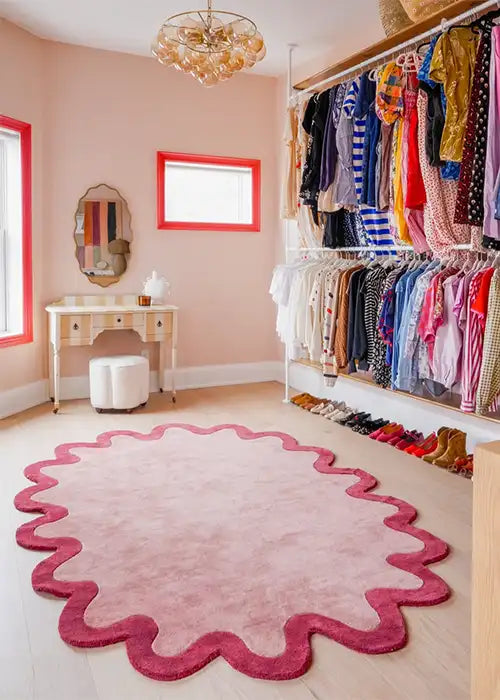 A beautifully curated dressing room with a pink and red scalloped-edge rug. The space features soft pink walls with coral window frames, an elegant vintage-style vanity with a round ottoman, and an open wardrobe displaying neatly hung clothing and organized shoes