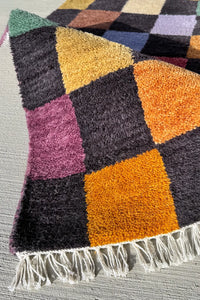 Handmade wool rug featuring a vibrant checkerboard pattern with mustard, burnt orange, sage, lilac, red, and blue squares.