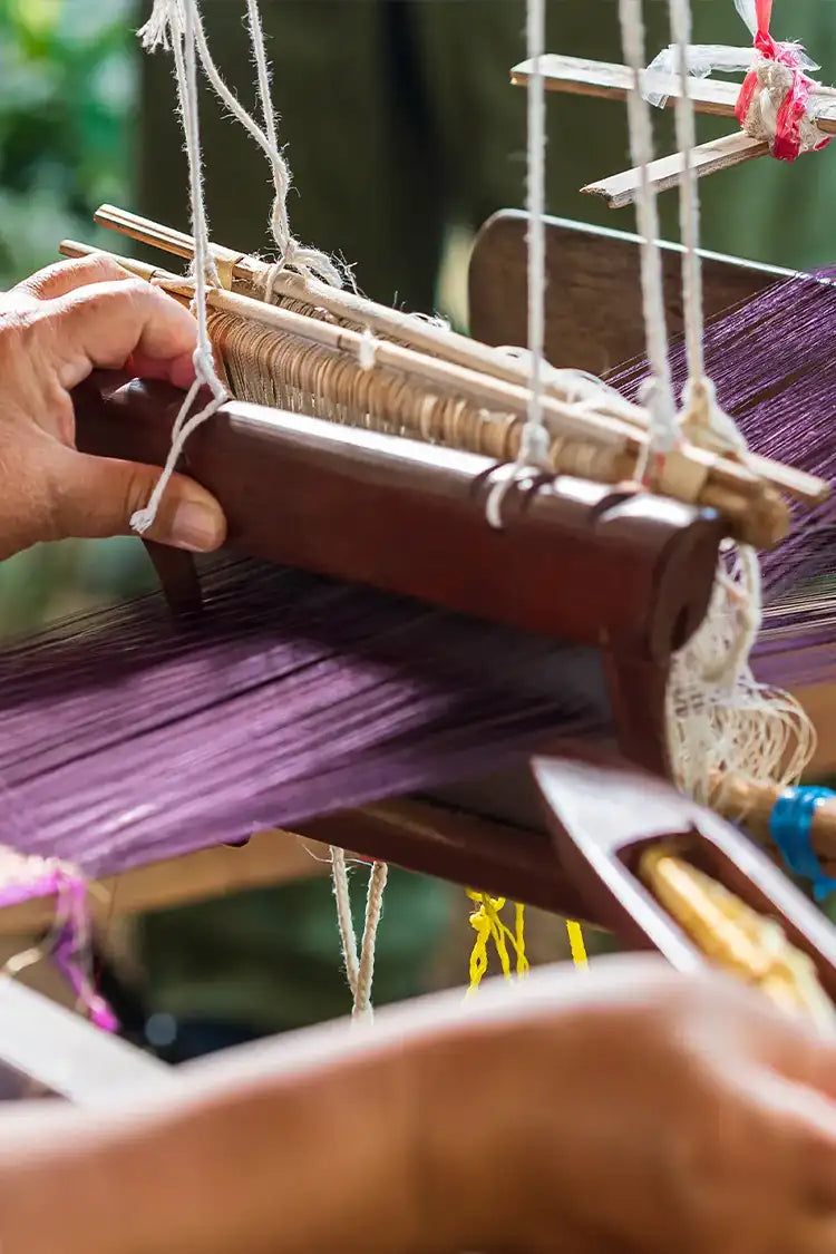 A close-up of an artisan’s hands working on a traditional wooden loom, weaving deep purple threads with precision and care.