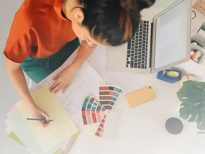 An interior designer working on a floor plan sketch with a set of color swatches, a laptop, and a phone on a bright workspace.