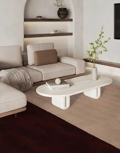 Modern neutral-toned living room with a cream sectional sofa, a sculptural white coffee table, and layered area rugs in beige and dark brown. Floating shelves with decor and a minimalist vase with greenery complete the space.