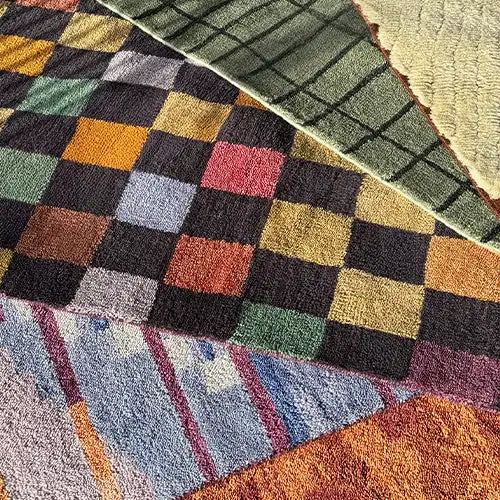 A collection of colorful, custom wool rugs featuring checkered, abstract, and geometric designs, laid out in natural sunlight.