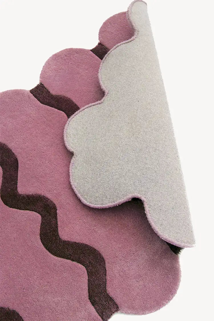Close-up of the Sculpted Edge Rug in soft pink and brown, showcasing its scalloped edges and layered design. The backing is partially flipped to reveal its durable construction.