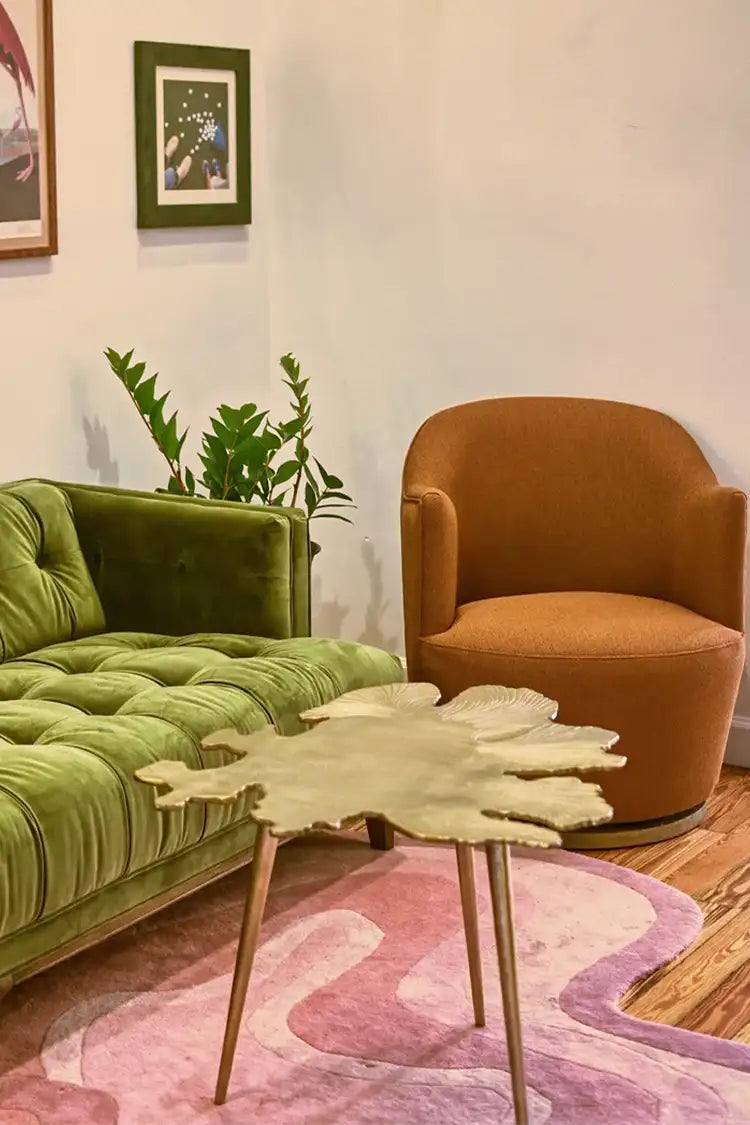 A cozy living room with a green velvet sofa, an amber armchair, and a pink modern rug, styled with vibrant and natural decor elements.