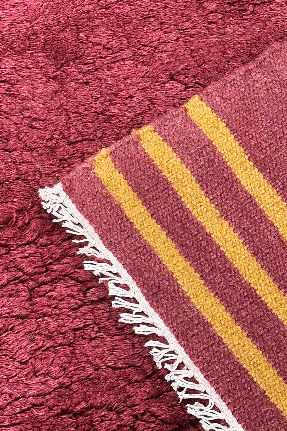 Corner of burgundy shag rug with mustard yellow stripes and white fringe, made from plush tufted wool