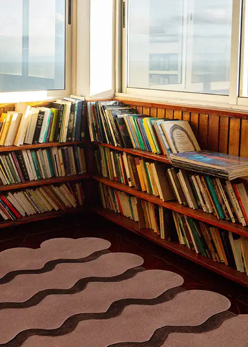 A cozy reading nook with a soft sculpted edge rug in neutral tones, complemented by a wall-to-wall wooden bookshelf and a scenic window view