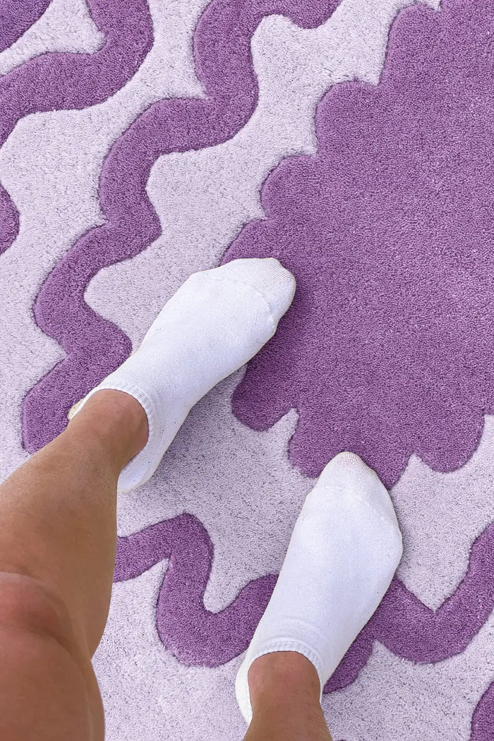 Feet standing on purple ripple oval wool rug with scalloped border and colorful design.