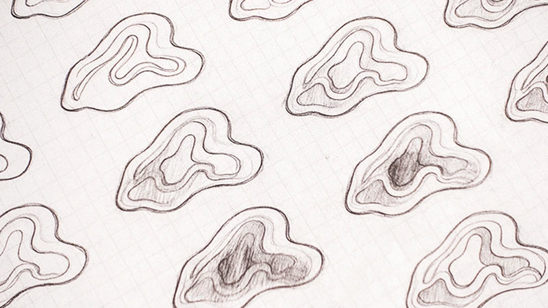 A close-up of a white fabric or paper surface with a repeating pattern of hand-drawn gray contour-like shapes outlined in dark gray.