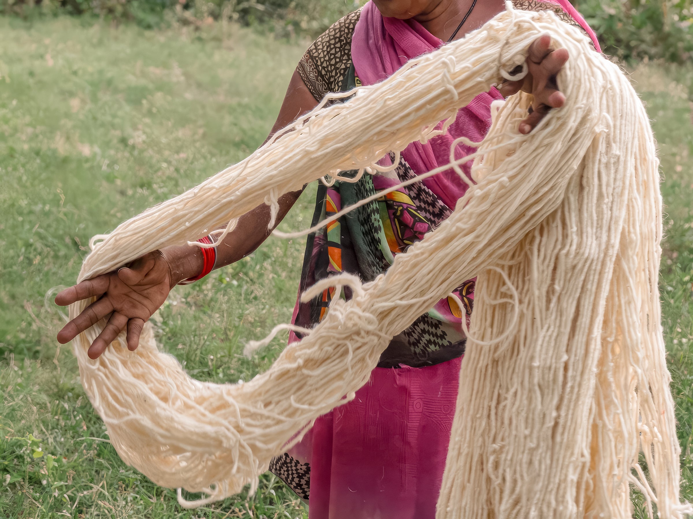 A person outdoors holds up a large bundle of thick, off-white yarn or thread. The person is wearing a pink and multicolored sari, and the background is a grassy field.