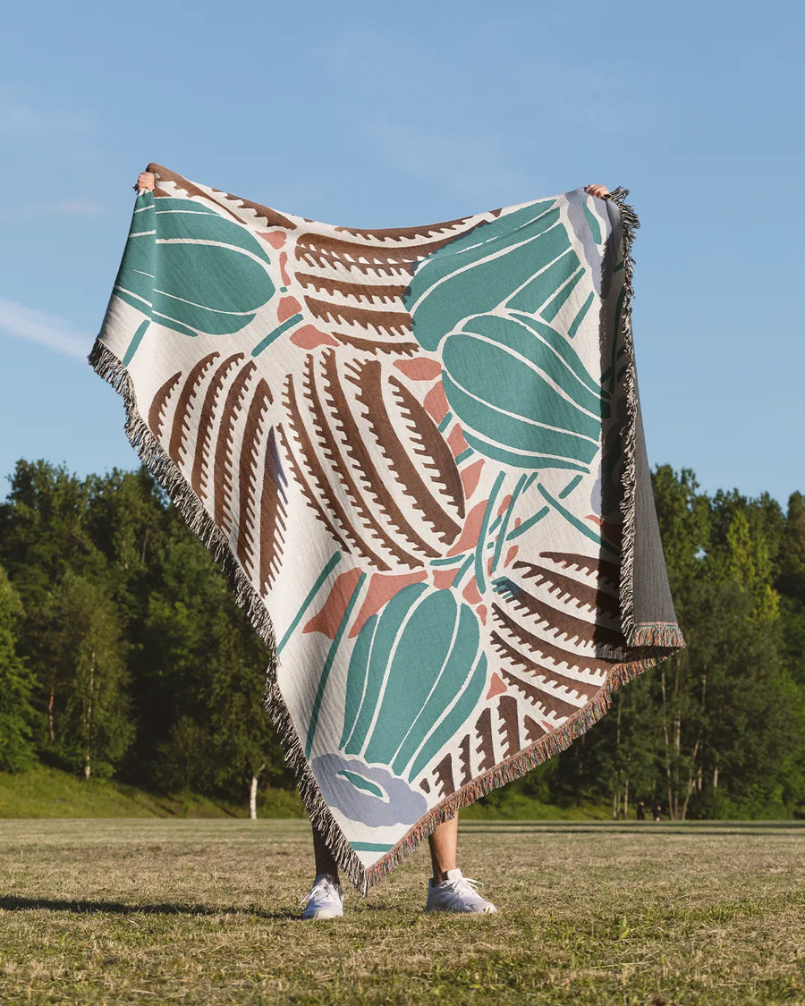 Desert-inspired woven throw blanket with cactus and floral patterns in earthy green, brown, and beige.
